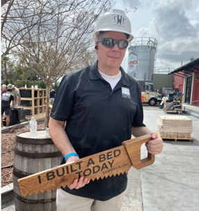 A man holding a wooden saw that says "I Built a Bed Today"